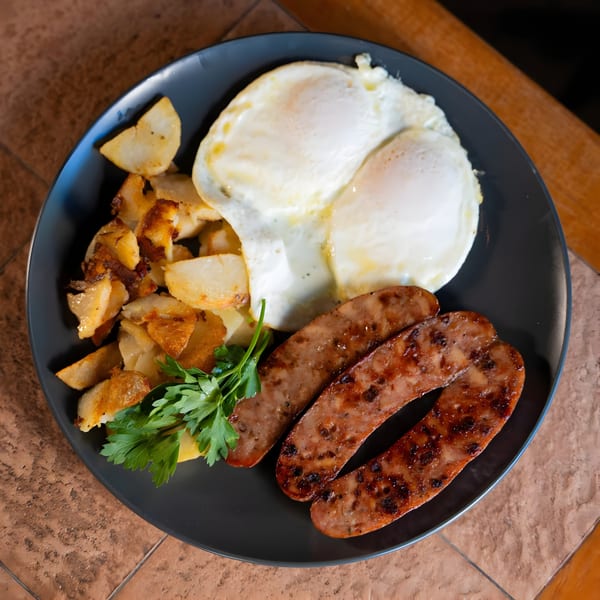 A breakfast plate with two fried eggs, crispy home fries, and two grilled sausage links, garnished with parsley.