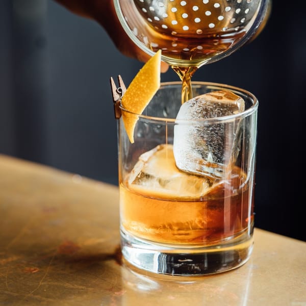A bartender pours a dark amber cocktail from a strainer into a glass with ice and an orange peel.