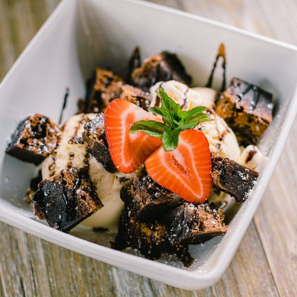 Brownie bites and vanilla ice cream dessert, topped with sliced strawberries and mint, in a white bowl.