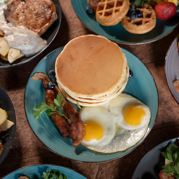 A breakfast plate with pancakes, fried eggs, sausage, bacon, and parsley on a blue plate.